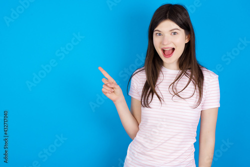 young beautiful Caucasian woman wearing stripped T-shirt over blue wall points aside on copy blank space. People promotion and advertising concept