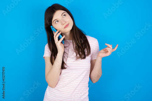 young beautiful Caucasian woman wearing stripped T-shirt over blue wall speaks on mobile phone spends free time indoors calls to friend.