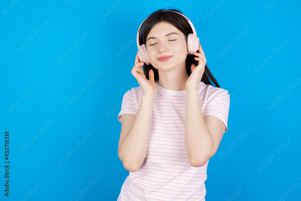 Pleased young beautiful Caucasian woman wearing stripped T-shirt over blue wall enjoys listening pleasant melody keeps hands on stereo headphones closes eyes. Spending free time with music Pleased young beautiful Caucasian woman wearing stripped T-shirt over blue wall enjoys listening pleasant melody keeps hands on stereo headphones closes eyes. Spending free time with music