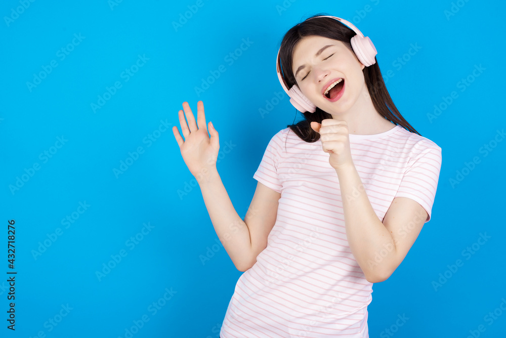 Happy young beautiful Caucasian woman wearing stripped T-shirt over blue wall sings favourite song keeps hand near mouth as if microphone wears wireless headphones, listens music Happy young beautiful Caucasian woman wearing stripped T-shirt over blue wall sings favourite song keeps hand near mouth as if microphone wears wireless headphones, listens music