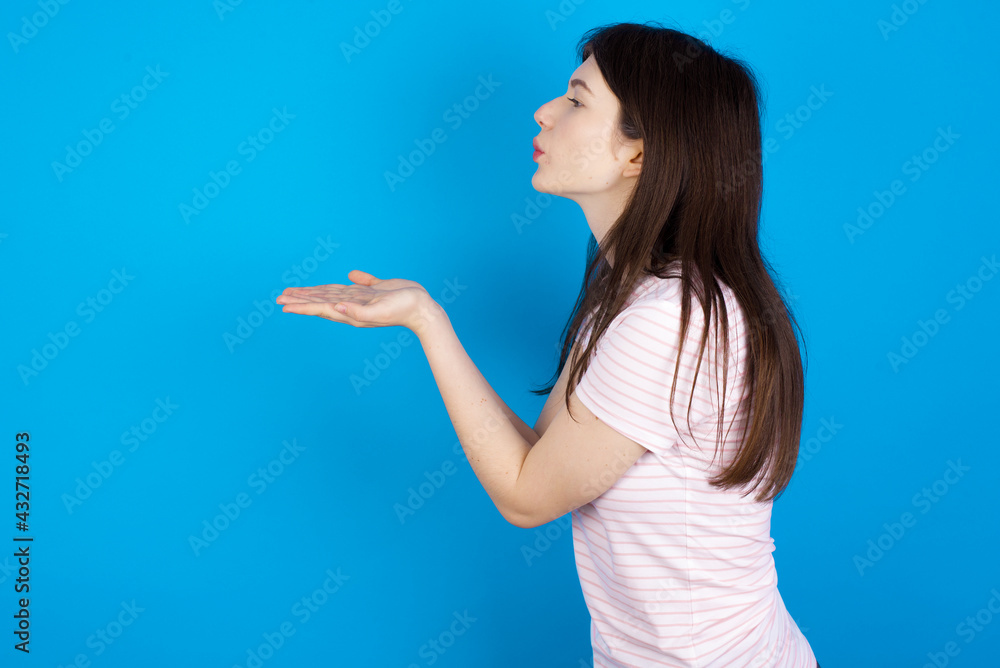 Profile side view view portrait of attractive young beautiful Caucasian woman wearing stripped T-shirt over blue wall sending air kiss Profile side view view portrait of attractive young beautiful Caucasian woman wearing stripped T-shirt over blue wall sending air kiss