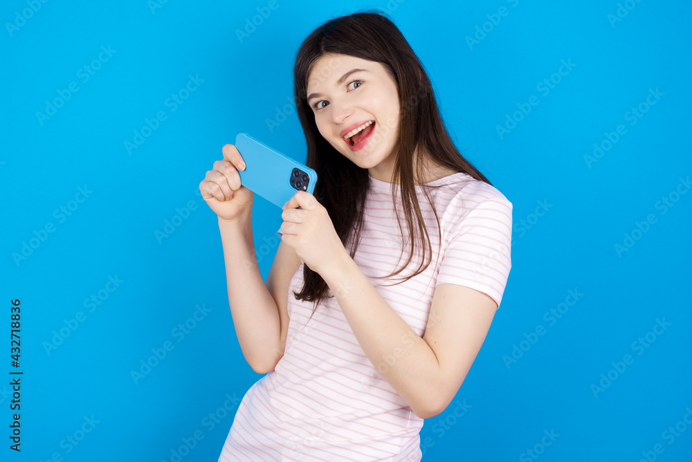 Nice addicted cheerful young beautiful Caucasian woman wearing stripped T-shirt over blue wall using gadget playing network game Nice addicted cheerful young beautiful Caucasian woman wearing stripped T-shirt over blue wall using gadget playing network game