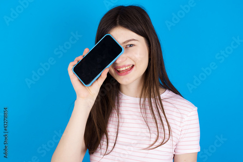 young beautiful Caucasian woman wearing stripped T-shirt over blue wall holding modern smartphone covering one eye while smiling