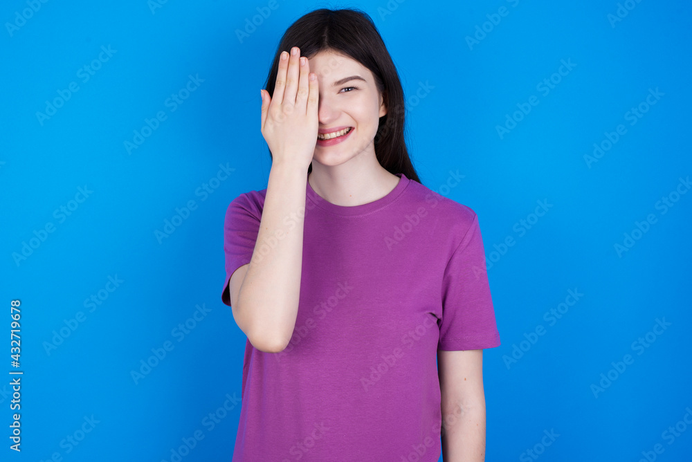 young beautiful Caucasian woman wearing purple T-shirt over blue wall covering one eye with her hand, confident smile on face and surprise emotion. young beautiful Caucasian woman wearing purple T-shirt over blue wall covering one eye with her hand, confident smile on face and surprise emotion.