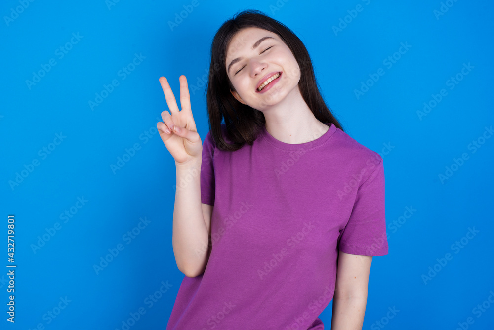 Fototapeta premium young beautiful Caucasian woman wearing purple T-shirt over blue wall smiling with happy face winking at the camera doing victory sign. Number two.