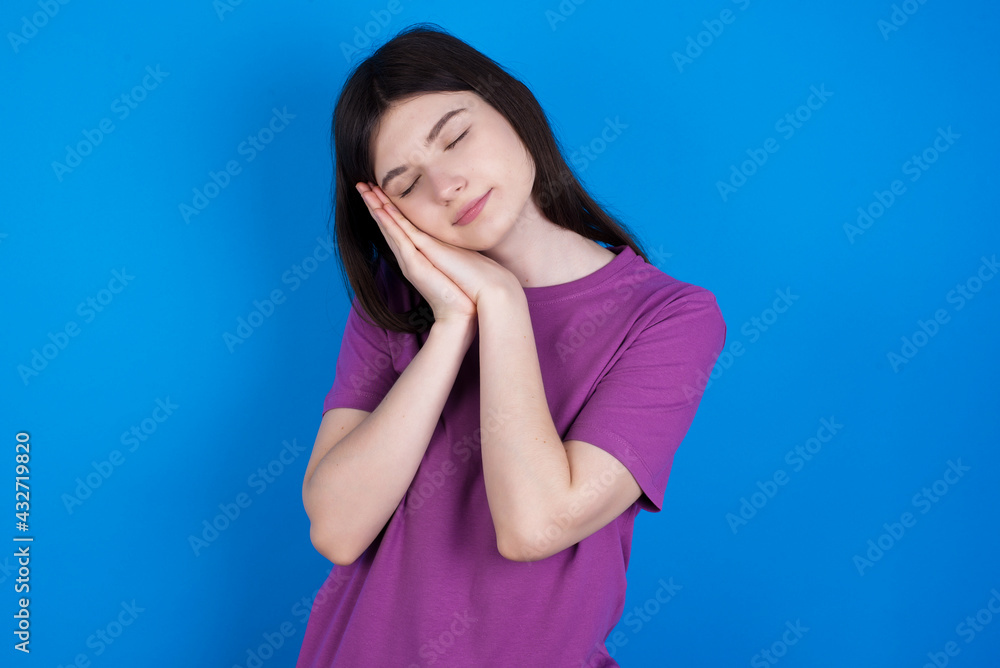 young beautiful Caucasian woman wearing purple T-shirt over blue wall sleeping tired dreaming and posing with hands together while smiling with closed eyes.