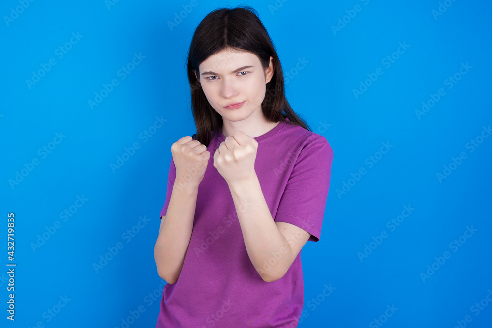 young beautiful Caucasian woman wearing purple T-shirt over blue wall Ready to fight with fist defense gesture, angry and upset face, afraid of problem. young beautiful Caucasian woman wearing purple T-shirt over blue wall Ready to fight with fist defense gesture, angry and upset face, afraid of problem.