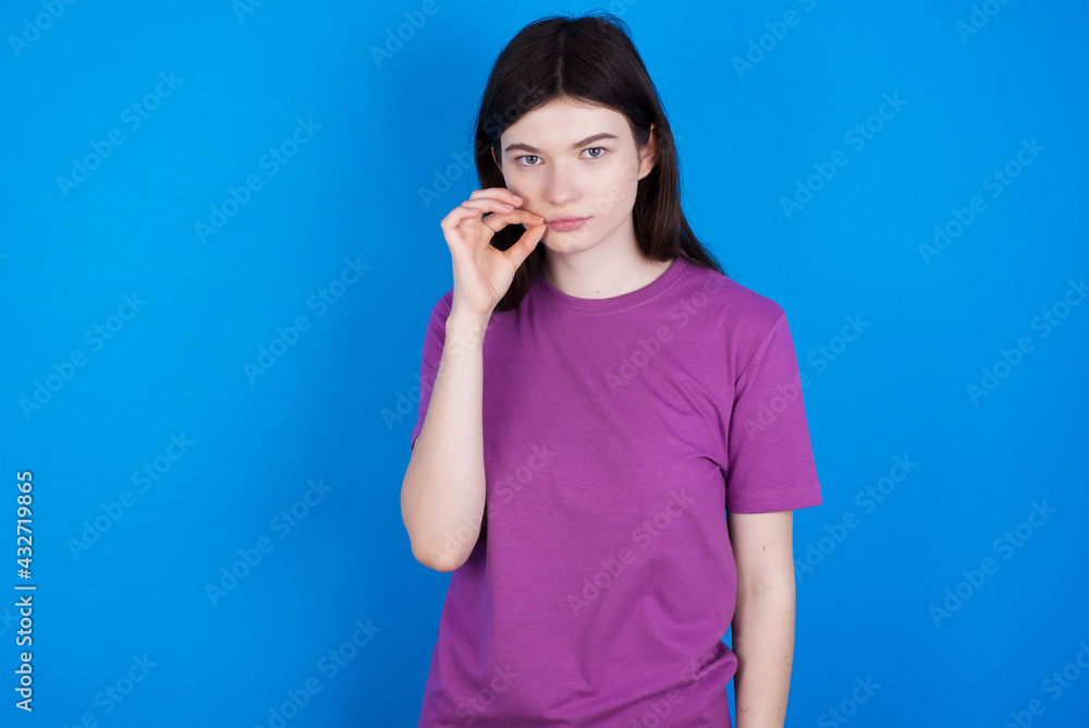 young beautiful Caucasian woman wearing purple T-shirt over blue wall mouth and lips shut as zip with fingers. Secret and silent, taboo talking. young beautiful Caucasian woman wearing purple T-shirt over blue wall mouth and lips shut as zip with fingers. Secret and silent, taboo talking.