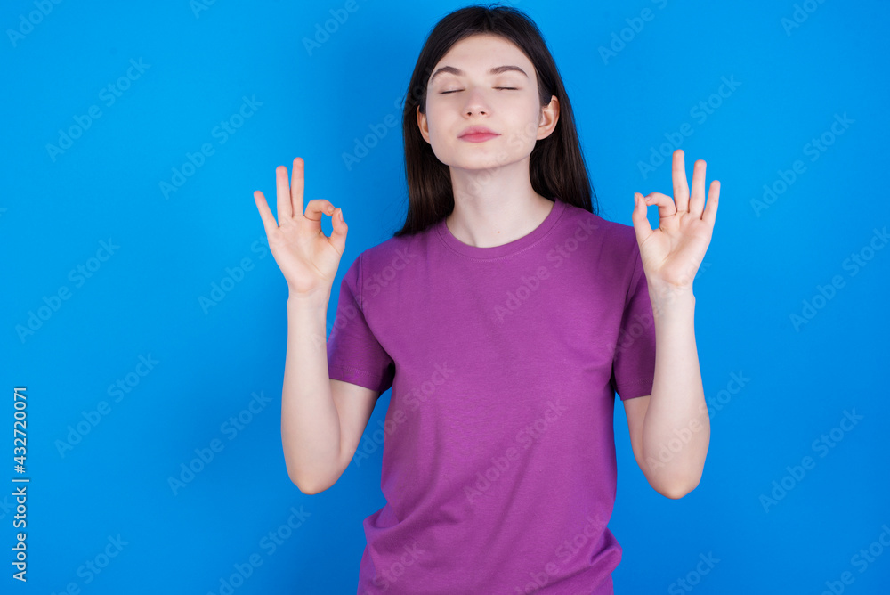 young beautiful Caucasian woman wearing purple T-shirt over blue wall relax and smiling with eyes closed doing meditation gesture with fingers. Yoga concept. young beautiful Caucasian woman wearing purple T-shirt over blue wall relax and smiling with eyes closed doing meditation gesture with fingers. Yoga concept.
