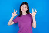 young beautiful Caucasian woman wearing purple T-shirt over blue wall showing and pointing up with fingers number eight while smiling confident and happy.