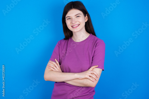 young beautiful Caucasian woman wearing purple T-shirt over blue wall happy face smiling with crossed arms looking at the camera. Positive person.