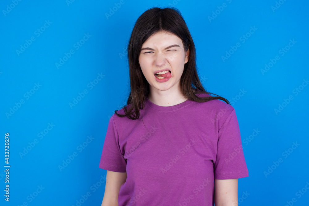 young beautiful Caucasian woman wearing purple T-shirt over blue wall sticking tongue out happy with funny expression. Emotion concept. young beautiful Caucasian woman wearing purple T-shirt over blue wall sticking tongue out happy with funny expression. Emotion concept.