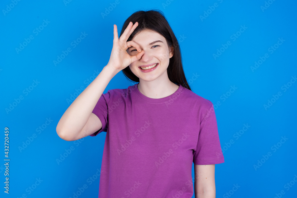 young beautiful Caucasian woman wearing purple T-shirt over blue wall doing ok gesture with hand smiling, eye looking through fingers with happy face. young beautiful Caucasian woman wearing purple T-shirt over blue wall doing ok gesture with hand smiling, eye looking through fingers with happy face.