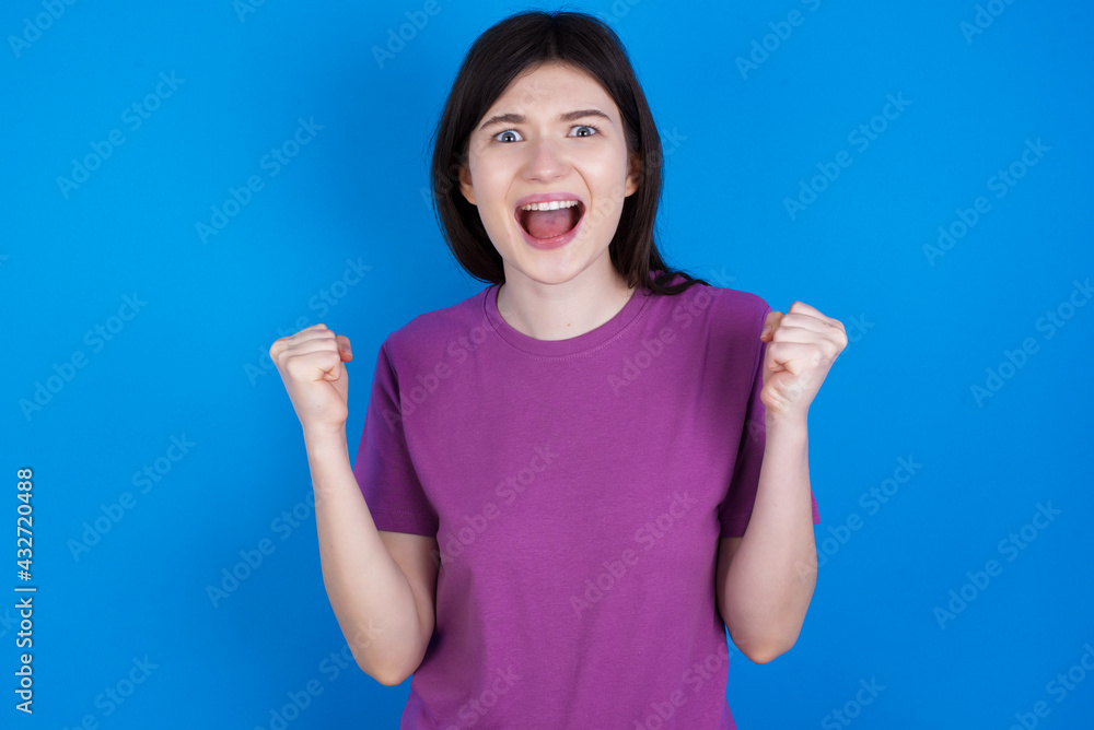 young beautiful Caucasian woman wearing purple T-shirt over blue wall celebrating surprised and amazed for success with arms raised and open eyes. Winner concept. young beautiful Caucasian woman wearing purple T-shirt over blue wall celebrating surprised and amazed for success with arms raised and open eyes. Winner concept.