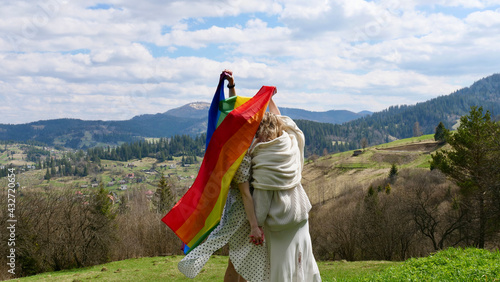 Lesbian brides, blonde women married in white wedding dresses with LGBT flag outdoors in the mountains. Homosexual girls with their backs holding each other by the ass without a face