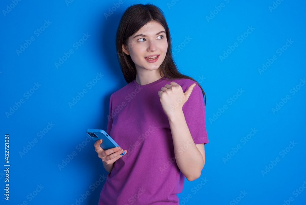 young beautiful Caucasian woman wearing purple T-shirt over blue wall using and texting with smartphone pointing and showing with thumb up to the side with happy face smiling young beautiful Caucasian woman wearing purple T-shirt over blue wall using and texting with smartphone pointing and showing with thumb up to the side with happy face smiling