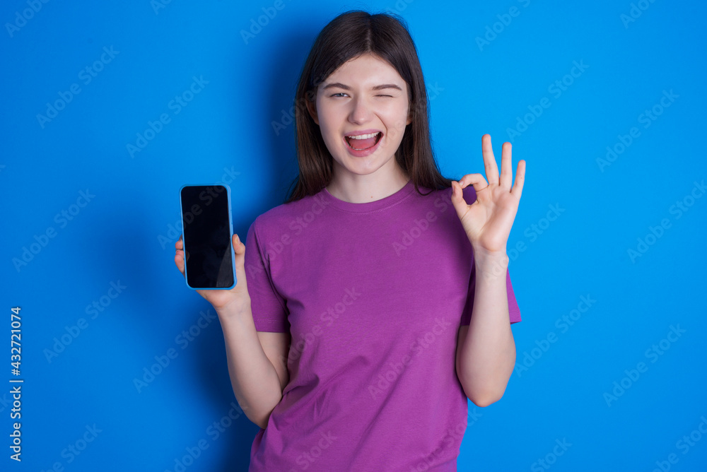 young beautiful Caucasian woman wearing purple T-shirt over blue wall holding in hands cell showing ok-sign young beautiful Caucasian woman wearing purple T-shirt over blue wall holding in hands cell showing ok-sign