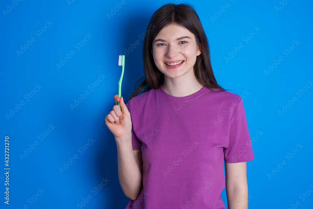 young beautiful Caucasian woman wearing purple T-shirt over blue wall holding a toothbrush and smiling. Dental healthcare concept. young beautiful Caucasian woman wearing purple T-shirt over blue wall holding a toothbrush and smiling. Dental healthcare concept.