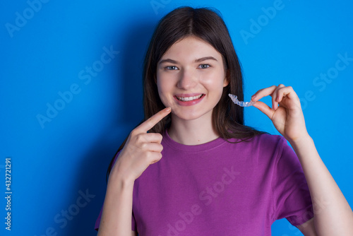 young beautiful Caucasian woman wearing purple T-shirt over blue wall holding an invisible aligner and pointing to her perfect straight teeth. Dental healthcare and confidence concept.