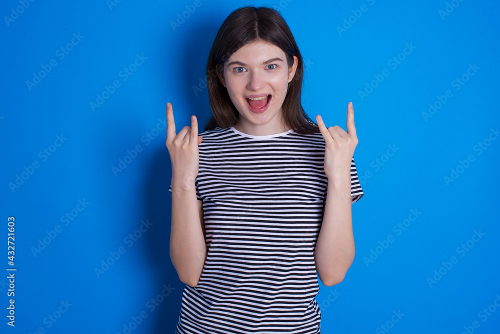 young beautiful Caucasian woman wearing pink jacket over blue wall makes rock n roll sign looks self confident and cheerful enjoys cool music at party. Body language concept. young beautiful Caucasian woman wearing pink jacket over blue wall makes rock n roll sign looks self confident and cheerful enjoys cool music at party. Body language concept.