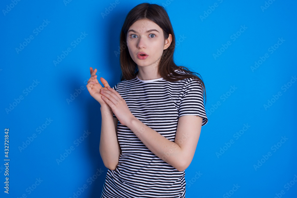 Fototapeta premium Surprised emotional young beautiful Caucasian woman wearing pink jacket over blue wall rubs palms and stares at camera with disbelief