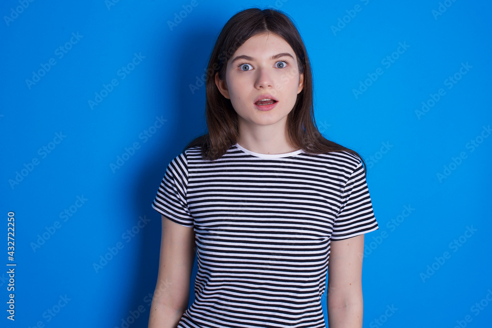 Shocked young beautiful Caucasian woman wearing stripped T-shirt over blue wall stares bugged eyes keeps mouth opened has surprised expression. Omg concept Shocked young beautiful Caucasian woman wearing stripped T-shirt over blue wall stares bugged eyes keeps mouth opened has surprised expression. Omg concept
