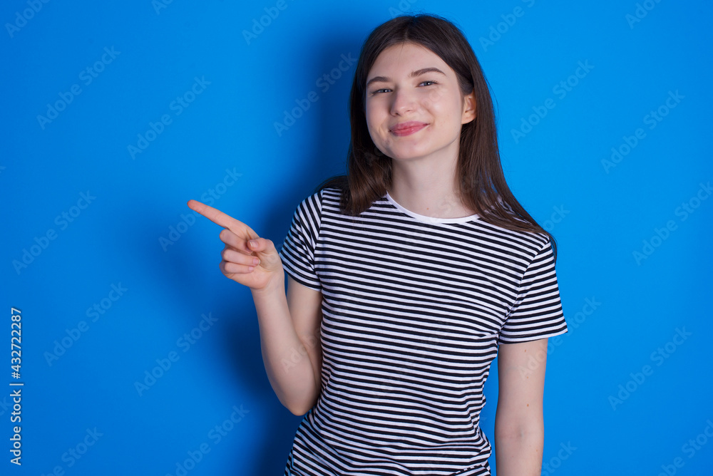 young beautiful Caucasian woman wearing stripped T-shirt over blue wall points to side on blank space demonstrates advertisement. People and promotion concept young beautiful Caucasian woman wearing stripped T-shirt over blue wall points to side on blank space demonstrates advertisement. People and promotion concept