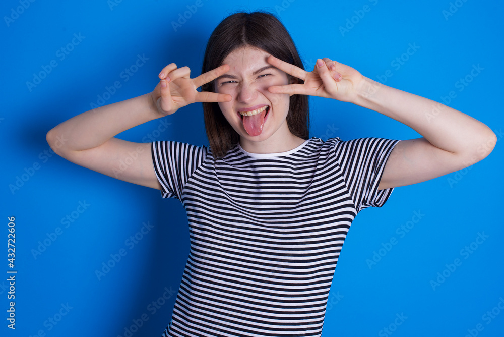 Cheerful positive young beautiful Caucasian woman wearing stripped T-shirt over blue wall shows v-sign near eyes open mouth Cheerful positive young beautiful Caucasian woman wearing stripped T-shirt over blue wall shows v-sign near eyes open mouth