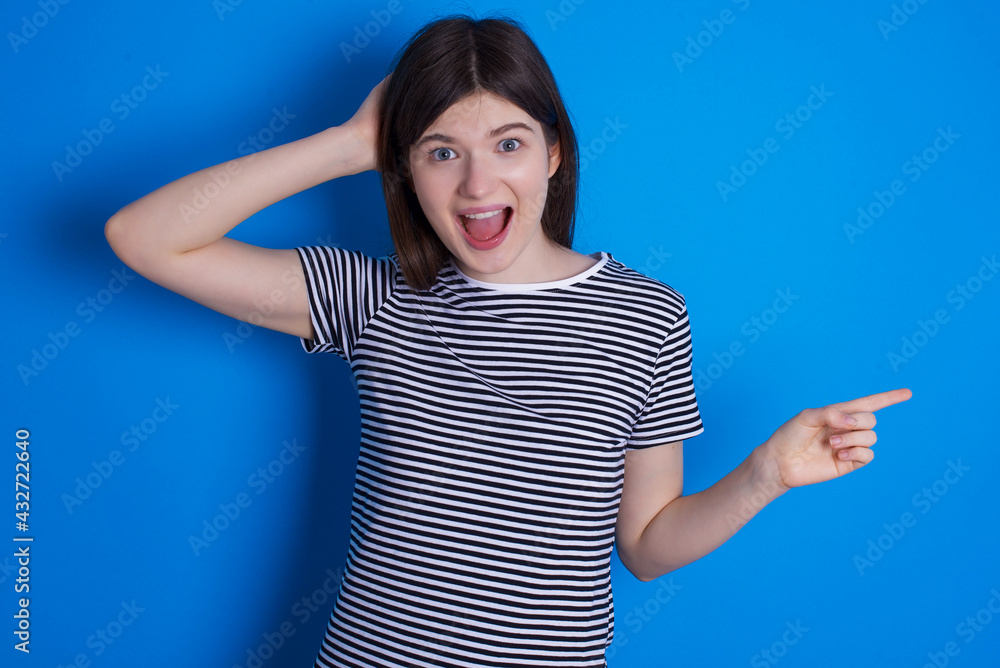 Surprised young beautiful Caucasian woman wearing stripped T-shirt over blue wall pointing at empty space holding hand on head Surprised young beautiful Caucasian woman wearing stripped T-shirt over blue wall pointing at empty space holding hand on head