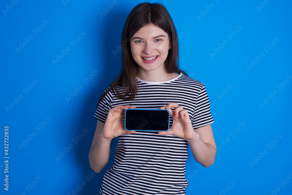 Cheerful cheery content young beautiful Caucasian woman wearing stripped T-shirt over blue wall holding in hands device hobby smm post blog Cheerful cheery content young beautiful Caucasian woman wearing stripped T-shirt over blue wall holding in hands device hobby smm post blog