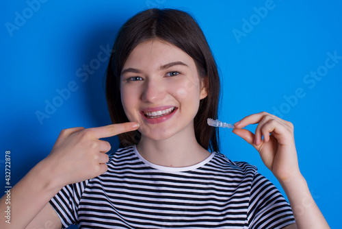 Fototapeta Naklejka Na Ścianę i Meble -  young beautiful Caucasian woman wearing stripped T-shirt over blue wall holding an invisible aligner and pointing to her perfect straight teeth. Dental healthcare and confidence concept.