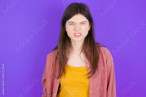 Mad crazy young beautiful Caucasian woman wearing casual clothes over purple wall clenches teeth angrily, being annoyed with coming noise. Negative feeling concept.