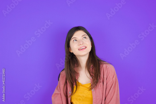 Portrait of mysterious young beautiful Caucasian woman wearing casual clothes over purple wall looking up with enigmatic smile. Advertisement concept.
