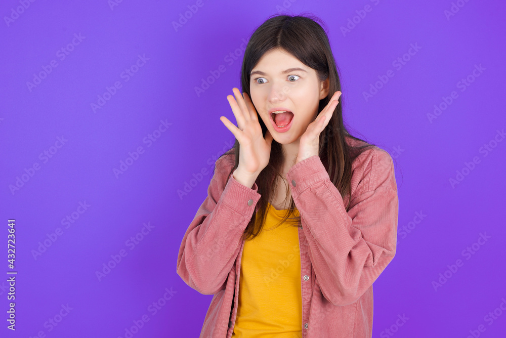 young beautiful Caucasian woman wearing casual clothes over purple wall with shocked facial expression, holding hands near face, screaming and looking sideways at something amazing. young beautiful Caucasian woman wearing casual clothes over purple wall with shocked facial expression, holding hands near face, screaming and looking sideways at something amazing.