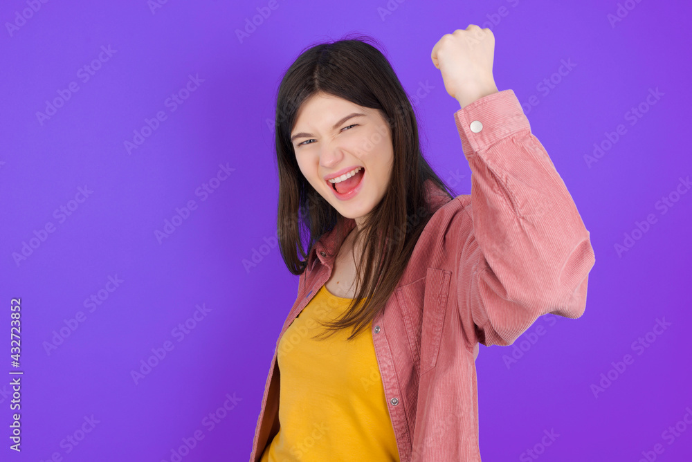 Overjoyed young beautiful Caucasian woman wearing casual clothes over purple wall glad to receive good news, clenching fist and making winning gesture. Overjoyed young beautiful Caucasian woman wearing casual clothes over purple wall glad to receive good news, clenching fist and making winning gesture.
