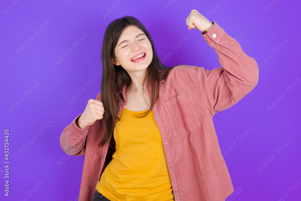 Attractive young beautiful Caucasian woman wearing casual clothes over purple wall celebrating a victory punching the air with his fists and a beaming toothy smile Attractive young beautiful Caucasian woman wearing casual clothes over purple wall celebrating a victory punching the air with his fists and a beaming toothy smile