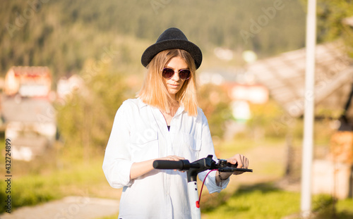 Young blond woman in black hat and sunglasses sets the ride mode on the touch panel. Stylish girl riding on electric scooter in city center. Summer leisure activity. Spending free time outdoors in