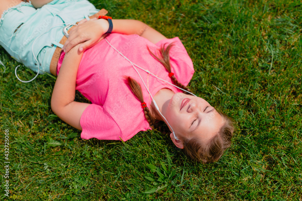 Caucasian girl in a pink T-shirt and white headphones listens to music. Girl student lies smiling on the green grass, turning her face to the sun's rays. Caucasian girl in a pink T-shirt and white headphones listens to music. Girl student lies smiling on the green grass, turning her face to the sun's rays.
