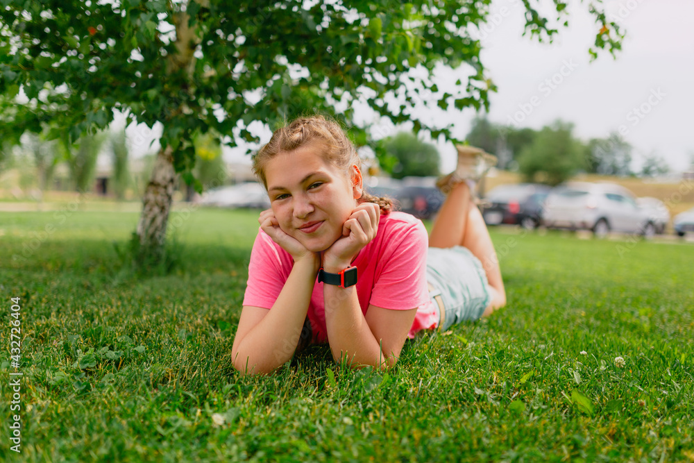 Fototapeta premium Caucasian girl with freckles and in a pink T-shirt lies on the grass. Girl student smiles and enjoys the beautiful summer weather.