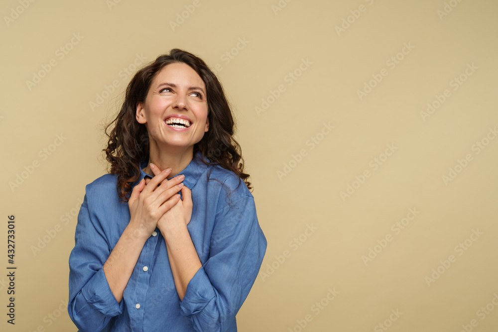 Excited casual young woman in jeans shirt with curly hair, happy smile look up to empty space. Joyful millennial female holding hands together under chin dreaming isolated on beige studio background Excited casual young woman in jeans shirt with curly hair, happy smile look up to empty space. Joyful millennial female holding hands together under chin dreaming isolated on beige studio background