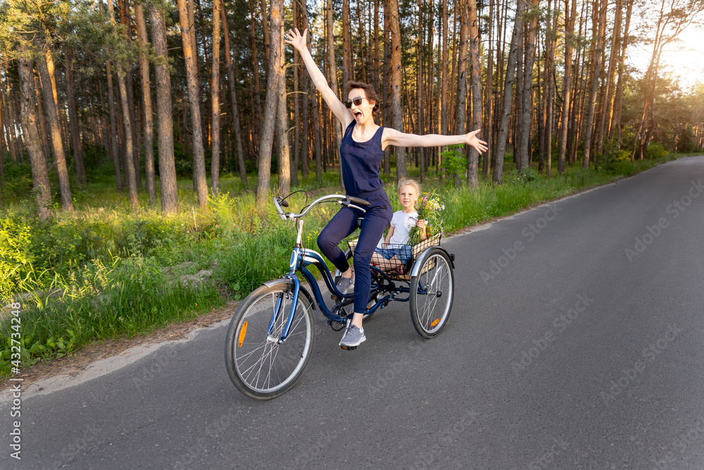 Young adult caucasian mom enjoy having leisure fun riding bicycle with cute adorable blond daughter holding wild field flower at scenic rural country road on bright sunny day. Countryside vacation