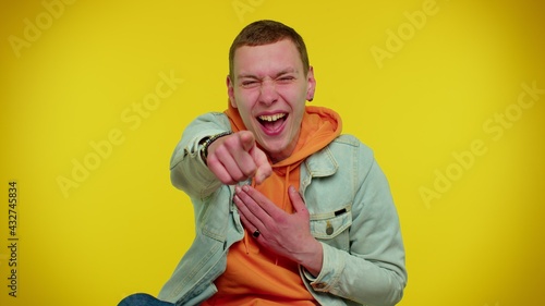 Amused teen stylish man in denim jacket pointing finger to camera, laughing out loud, taunting making fun of ridiculous appearance, funny joke. Young guy boy posing on yellow studio wall background © Andrii Iemelianenko