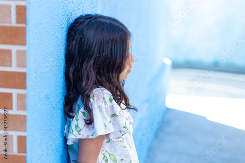 Uma criança, menina, brasileira dos cabelos pretos e lisos, de perfil, encostada em parede com expressão triste. photo