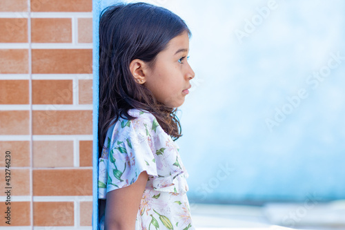 Uma criança, menina, brasileira dos cabelos pretos e lisos, de perfil, encostada em parede com expressão triste. photo