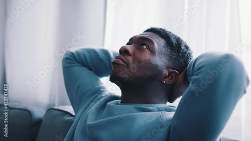 Slow-motion footage of a serious man thinking while sitting on a blue sofa. Hands behind the head and looking at the ceiling of the room. Focused. Cozy room with white curtains in the background.  photo