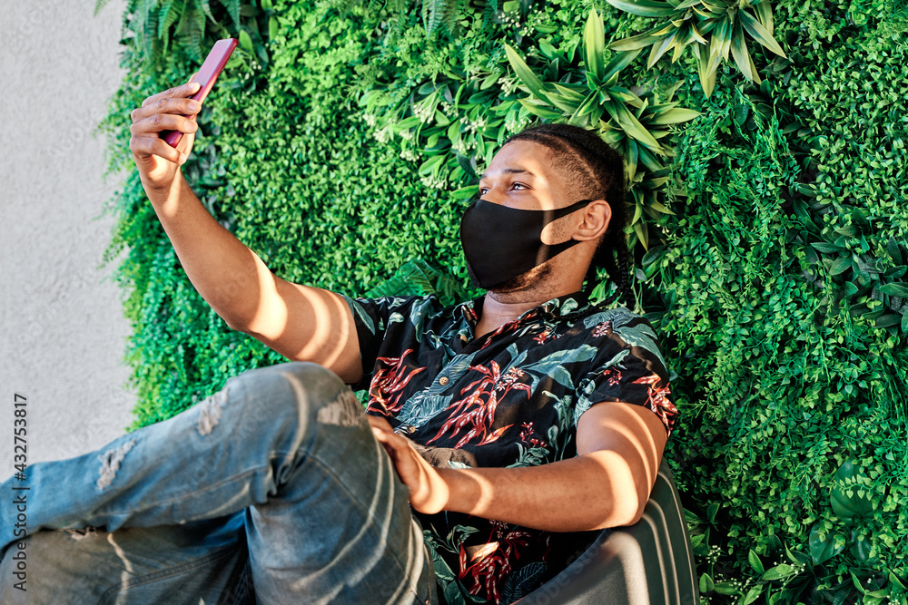 young latin man sitting in a naturally decorated environment taking a selfie and wearing a mask