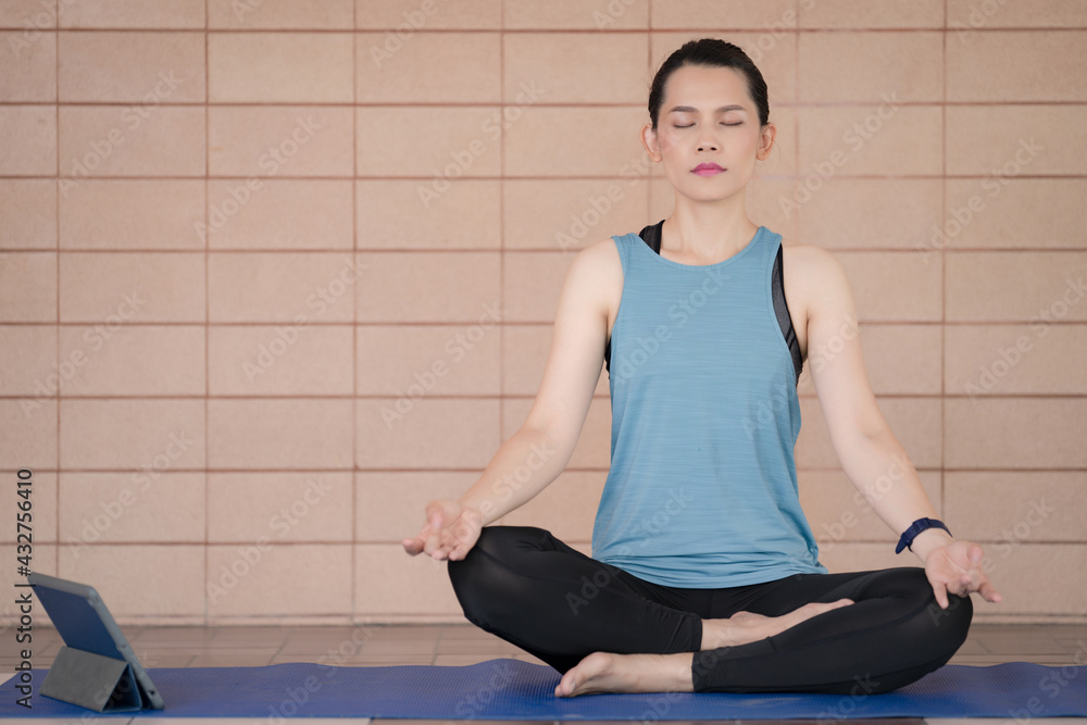 A middle-aged calm woman in relaxed sports outfits sits in a lotus position practicing yoga and meditation with relaxed eyes closed while listening to spiritual practices lesson on a tablet at home A middle-aged calm woman in relaxed sports outfits sits in a lotus position practicing yoga and meditation with relaxed eyes closed while listening to spiritual practices lesson on a tablet at home