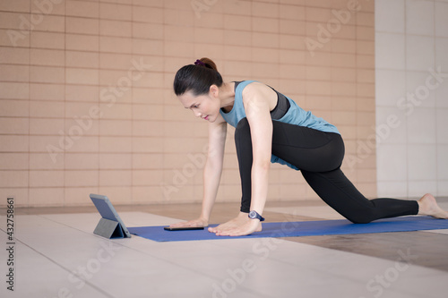 A middle-aged Asian woman in relaxed sports outfits doing an exercise workout training program follows an online workout lesson on a tablet at home during the COVID-19 pandemic and city lockdown