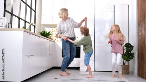 Excited mom or babysiter and two kids boy and girl jumping, dancing laughing in modern scandinavian house kitchen. Happy family mother daughter son having fun enjoy playing singing together at home.