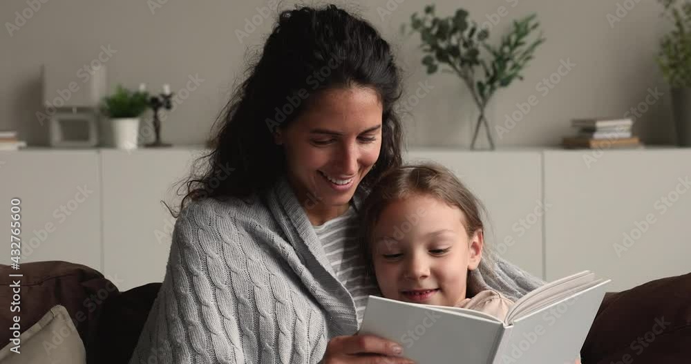 Head shot happy bonding young mother resting on sofa covered in warm plaid with cute little 7s daughter, enjoying reading paper book with fairy tale, spending peaceful carefree weekend time at home. Head shot happy bonding young mother resting on sofa covered in warm plaid with cute little 7s daughter, enjoying reading paper book with fairy tale, spending peaceful carefree weekend time at home.